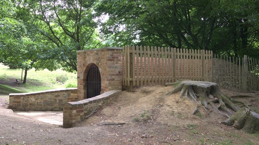 A sideview of a brick fronted structure with an arched entranceway. There is a black metal gate across the entrance and a fence running down the side of the structure. There are trees in the background.